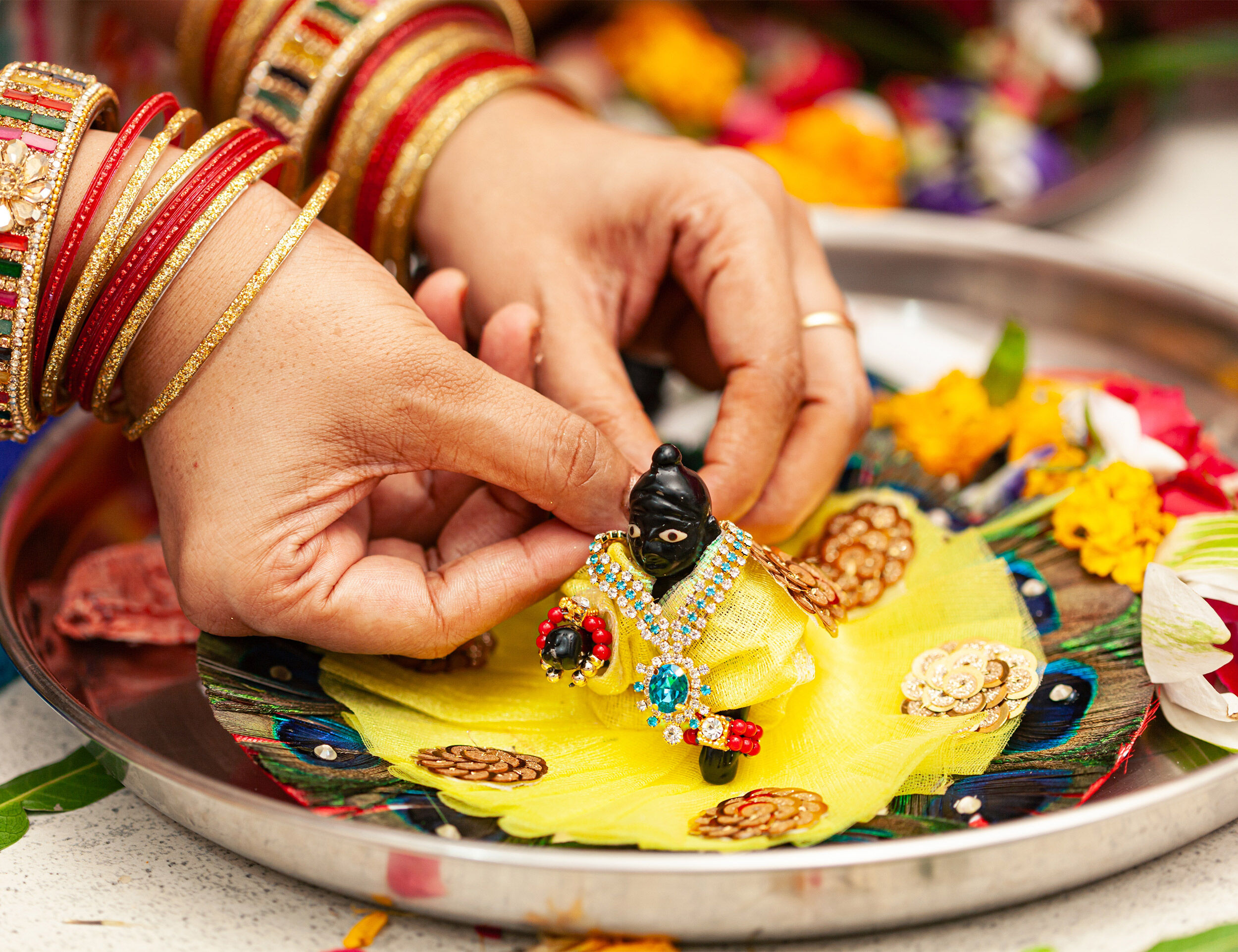 Woman adorning a small Lord Krishna idol with yellow traditional attire and jewelry during Krishna Janmashtami rituals, surrounded by flowers and peacock feathers